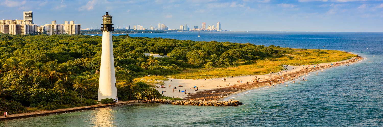 Aerial helicopter photograph of the historic Cape Florida Lighthouse in Key Biscayne, Florida, surrounded by sandy beaches, coastal vegetation, and Atlantic Ocean views.