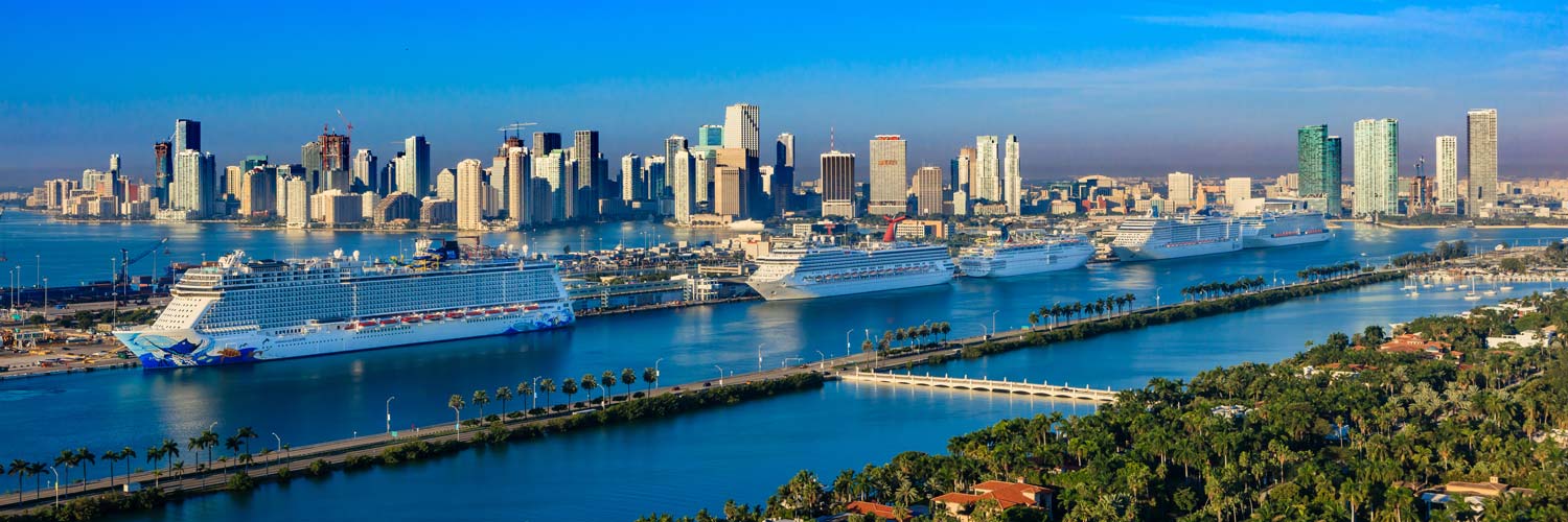 Aerial helicopter view of PortMiami featuring cruise ships, waterfront terminals, Biscayne Bay, and the Downtown Miami skyline.